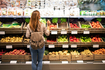 Good looking woman standing in front of vegetable shelves choosing what to buy. Buying groceries and healthy organic food in supermarket. - obrazy, fototapety, plakaty