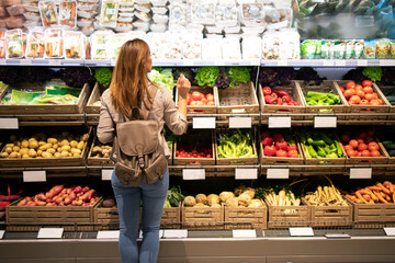 Good looking woman standing in front of vegetable shelves choosing what to buy. Buying groceries and healthy organic food in supermarket.
