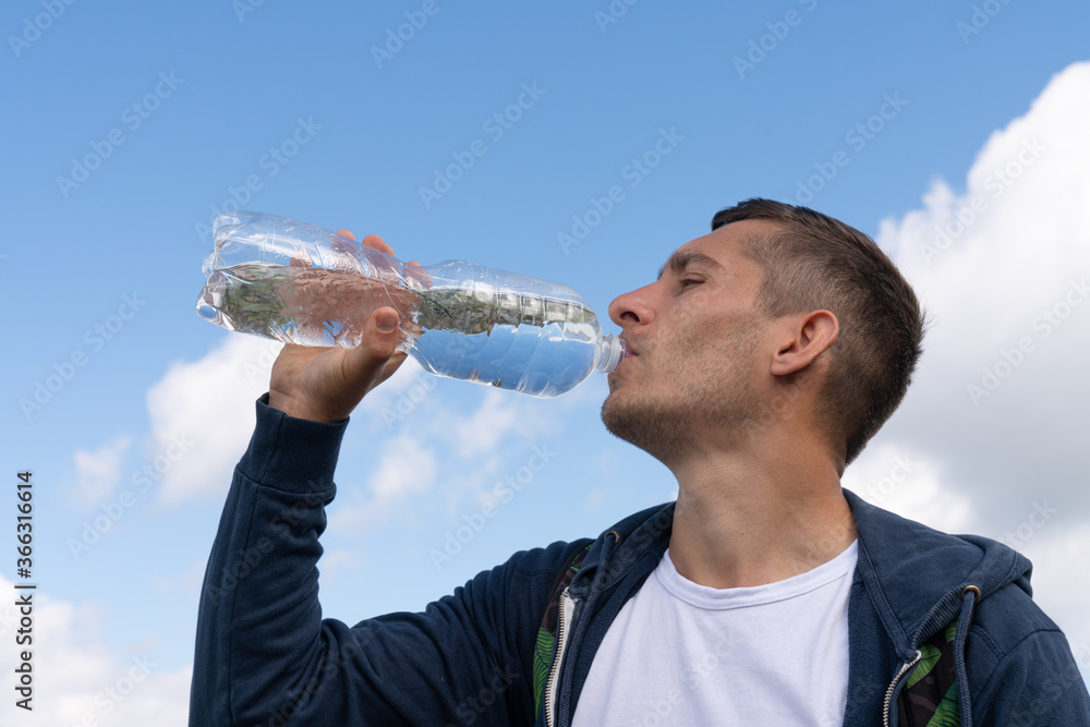 Wall mural Young sporty man drinking water from bottle against blue sky on sunny day