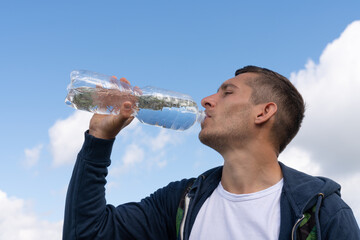 Young sporty man drinking water from bottle against blue sky on sunny day