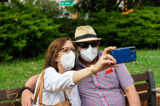 Photo Of A Middle Age Couple Sitting On A Bench Taking A Selfie And Wearing White Face Masks In The Park