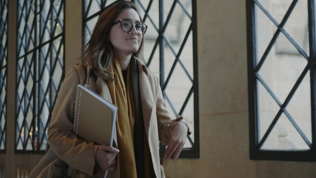 Female Student Walking In University Hallway. Woamn Holding Notebook In Hand