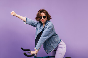 Funny girl in casual clothes posing on bike. Studio portrait of carefree curly lady sitting on bicycle.