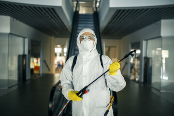 Portrait of a man in a sanitizing disifection suit holding spray near the escalator in an empty...