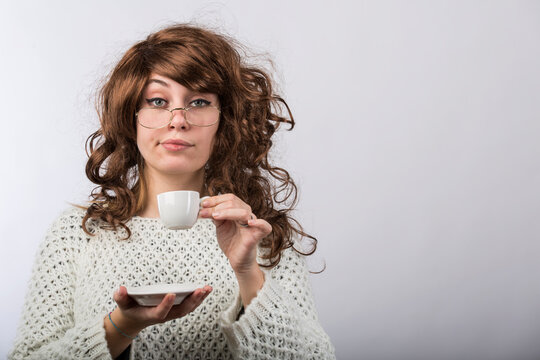 Signora con capelli mossi castani e maglione bianco beve il suo caffe da una tazzina , isolata su sfondo bianco 