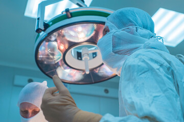 close-up of surgeons in a sterile mask, against the background of an operating lamp, with raised hands, the surgeon's assistant looks at the indicators in a monitor, sterile gloves and a mask.