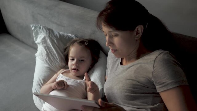 Mom and little daughter are lying in bed with wake cartoon on a tablet. mother and baby make video call to father. Technology concept, new generation, family, connection, parenthood