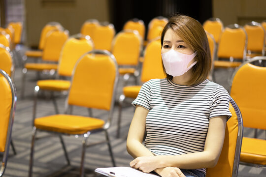 An Asian woman with a golden-bronze hair wearing a striped shirt wear facemark sitting and waiting for something in a chair amid an empty chair in yellow.