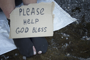 Feet of a homeless man sitting on the street in torn socks and a sign with the inscription....