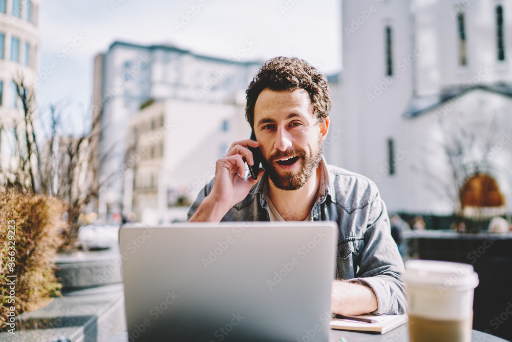 Poster portrait of positive bearded young man smiling at camera while calling on smartphone during remote w