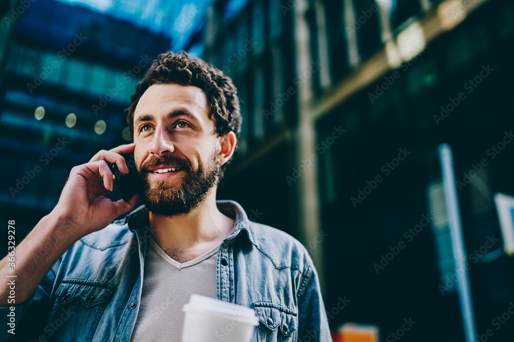 Poster bearded young man laughing while calling on smartphone device strolling in urban setting of modern c
