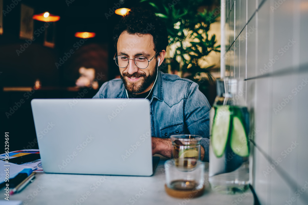 Poster positive bearded hipster student watching funny tutorial online in earphones connected to laptop dev