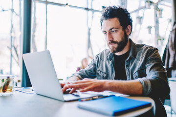 Fototapeta premium Concentrated bearded young man searching information on website and working on freelance on development graphic project at modern laptop using wireless internet connection in cafe interior