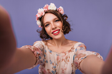 Elegant white girl in circlet of flowers making selfie and smiling. Studio portrait of relaxed inspired lady with short haircut.