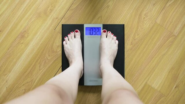 A woman gets on the scales and sees her weight at 103.5 lb. The girl is cunning and pushes off the floor to reduce the figure. First-person view. Comic image of a losing weight woman