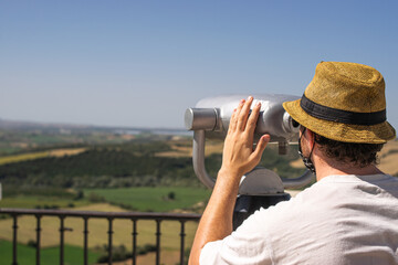 Photo of a young and attractive man wearing a reusable facce mask looking to the nature through some binoculars in a view point. Tourism during coronavirus outbreak