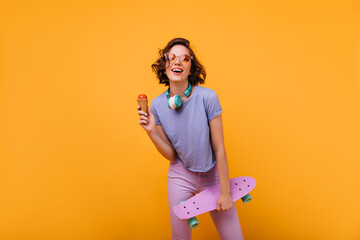 Magnificent white girl in casual clothes posing in headphones. Studio portrait of beautiful lady with skateboard eating ice cream on orange background.