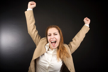 ragazza bionda con giacca beige esulta felice tirando i pugni in alto, isolata su sfondo nero