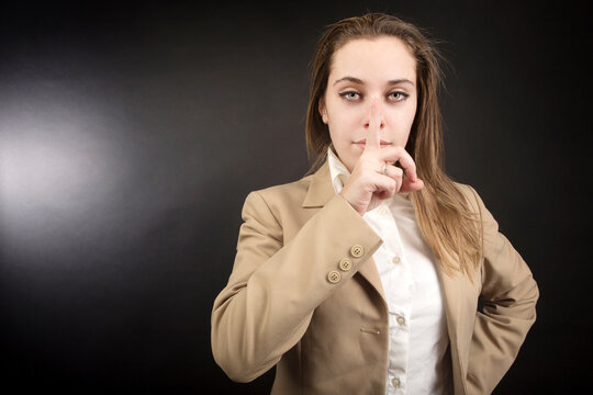 ragazza bionda con giacca beige fa il gesto del silenzio portando l'indice al naso, isolata su sfondo nero