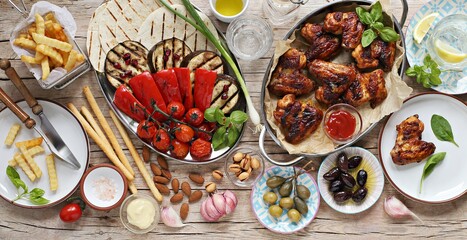 Chicken wings, roasted vegetables, grilled tortilla and appetizers variety serving on party outdoor table. Mediterranean dinner table concept. Overhead view.