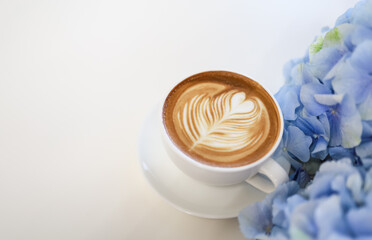 Closeup of white cup of hot coffee latte with milk foam heart shape art on white table with blue and purple hydrangea flower.