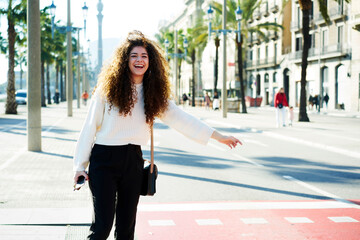 Cheerful casually dressed hipster girl with curly hair having fun while stopping taxi standing on city avenue, funny female traveler enjoying sunny weather and weekend trip hailing cab on road