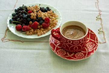 A red ceramic cup with freshly brewed coffee and a plate with fresh blueberries, red and black raspberries, cherries and white currants.