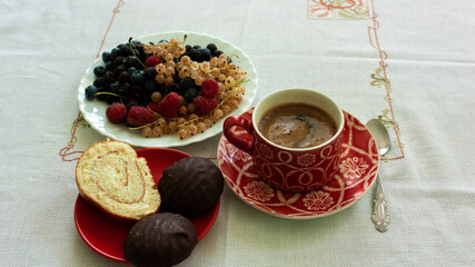 A plate with fresh blueberries, red and black raspberries, cherries and white currants, a red ceramic cup with freshly brewed coffee and a plate with a sweet dessert.