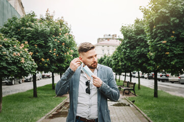 Stylish presentable Caucasian man puts on a disposable medical mask on the street. Protection during the coronavirus.