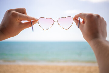 I like holidays. hands hold pink heart-shaped sunglasses. in the background a sandy beach and blue...