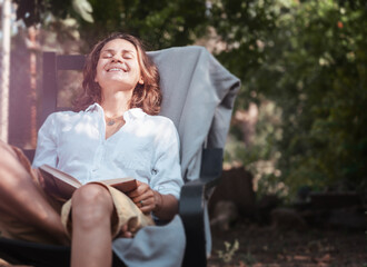 Naklejka premium Young beautiful girl woman enjoys nature and reading a book in the summer in the garden of her house sitting on a chair