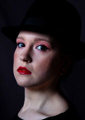 Young woman in heavy make up wearing a black hat. In low key lighting.