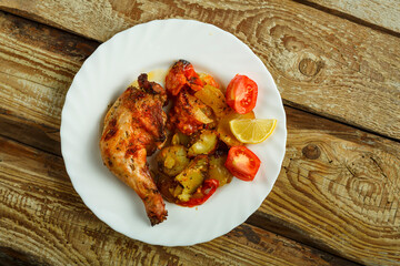 Baked chicken leg with potatoes and tomato on a white plate on a wooden table.