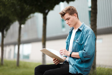 Young student sitting on a bench near the university building with a clipboard in hand preparing...
