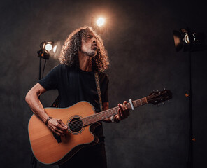 Middle aged hispanic musician in black t-shirt emotionally singing and playing guitar. View of musician in the spotlight