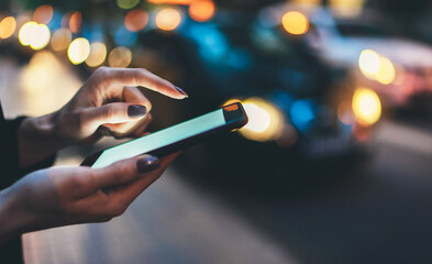 Female hands pointing on screen smartphone background headlights auto in night city street, tourist girl using internet technology calls a taxi standing next to the road