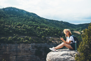 Naklejka na ściany i meble girl traveler in hat resting during vacation in nature and plans route online map in smartphone, tourist uses mobile phone on background of panorama mountain landscape, empty space