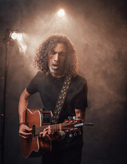 Hispanic musician in black t-shirt emotionally singing and playing guitar in stage lights, surrounded by smoke