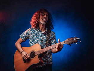 Obraz premium Happy middle aged hispanic musician singing and playing guitar on a dark illuminated by blue and red light. Concept of music, hobby, festival