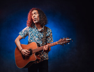 Obraz premium Middle aged hispanic man musician in a hawaiian shirt playing guitar on a dark illuminated by blue and red light. Concept of music, hobby, festival