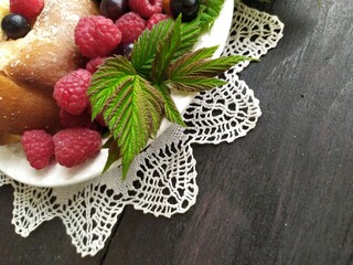 Pie with berries red ripe raspberries black currant green leaves on a white plate on a lace napkin on a black background