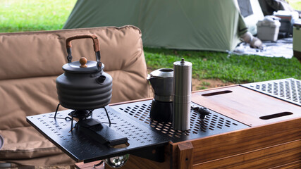 Vintage outdoor black kettle and coffee grinder on camping table with armchair on green lawn in...