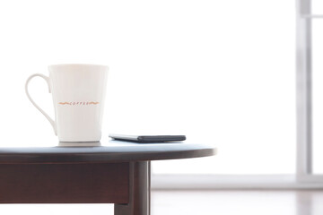 Close up white ceramic coffee cup and smart phone on oval wooden table with blurred frosted glass sliding door in white living room 