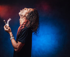 Middle aged hispanic male with long curly hair smoking from bong on a dark background illuminated...
