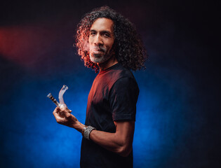 Middle aged hispanic male with long curly hair smoking from bong on a dark background illuminated by blue light