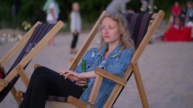 Young sleeply woman sits on the beach chair and watches environment. Beautiful female holds her drink and waits for sunset.