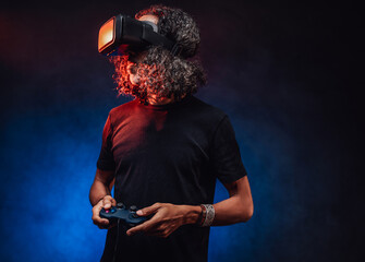 Middle aged hispanic male with long curly hair playing with virtual reality glasses and video game controller on a dark background illuminated blue light. Hi-tech concept with vr glasses