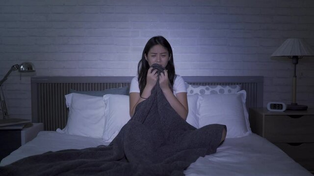asian lady sitting alone in dark bedroom seeing horror movie. girl scared by gruesome death scene and pulling up blanket, afraid to watch. real moments