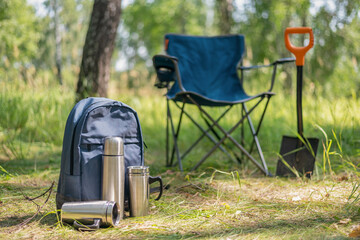 Backpack, vacuum flask, touristic chair and steel cup on the ground on the green forest background.