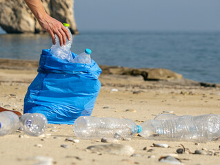 Hand picking up plastic bottles into blue trash bag. Cleaning on the beach.